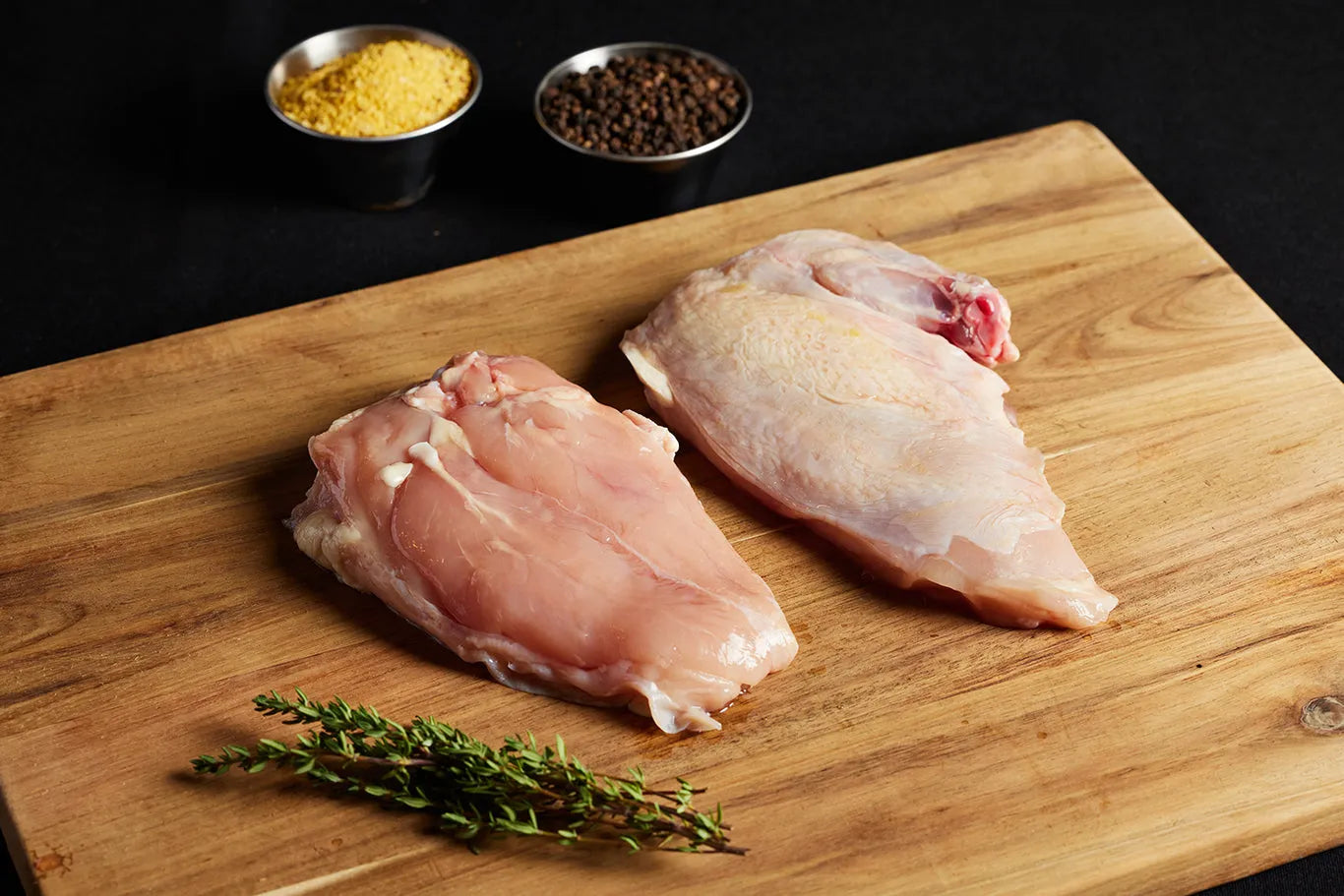 Two raw airline chicken breasts placed on a cutting board, accompanied by an assortment of spices.