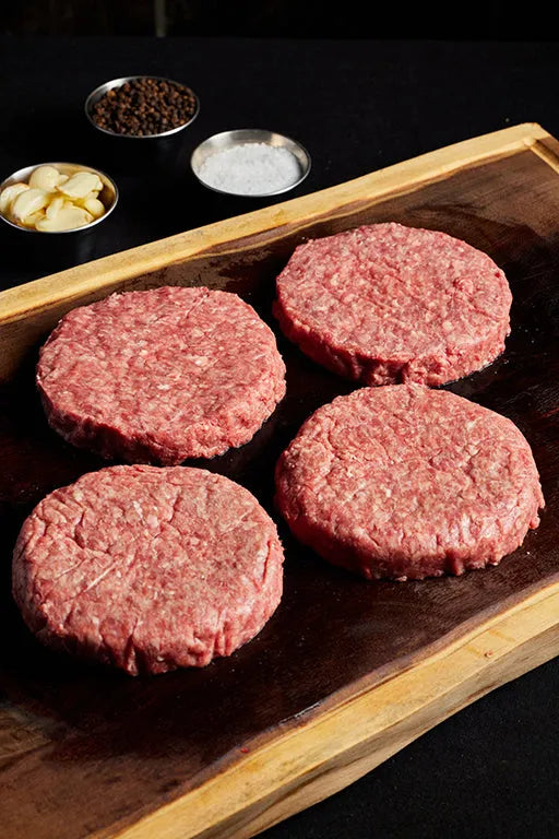 Four hamburger patties arranged on a wooden cutting board, ready for grilling and assembling into burgers.