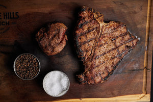 A T-Bone and Filet Mignon steak on a wooden cutting board, with spices ready to be added.