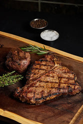 A T-Bone and Filet Mignon steak on a wooden cutting board, garnished with fresh herbs and spices.