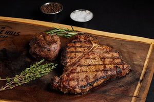 A T-Bone and Filet Mignon steak on a wooden cutting board, garnished with fresh herbs and spices for preparation.