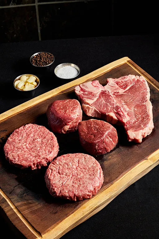 A cutting board displaying a T-bone steak, burgers, and filet mignons, accompanied by a spices for seasoning.