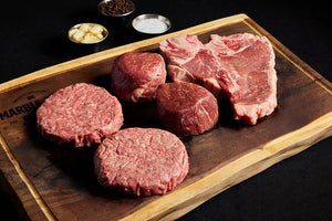 A cutting board displaying a T-bone steak, burgers, and filet mignons, accompanied by a spices for seasoning.