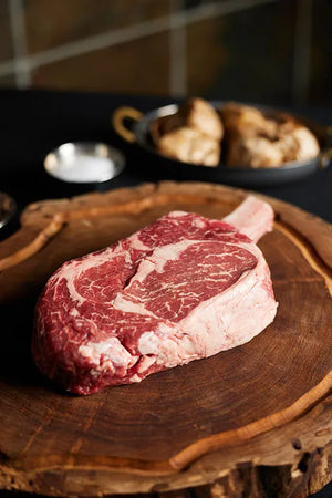 A Cowboy Ribeye steak resting on a wooden cutting board, showcasing its marbling and color.