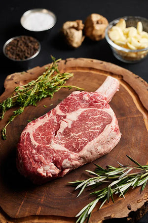 A Cowboy Ribeye steak rests on a wooden cutting board, surrounded by fresh herbs and an array of spices.