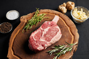 A Cowboy Ribeye steak rests on a wooden cutting board, surrounded by fresh herbs and an array of spices.