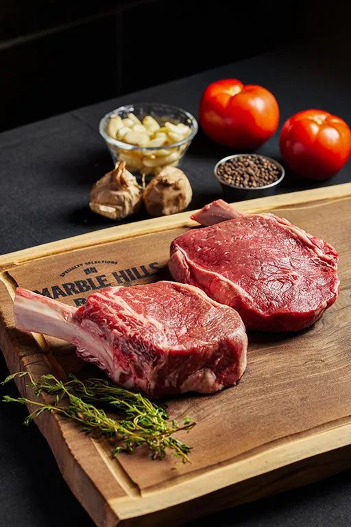 Two Cowgirl Ribeye steaks on a cutting board, accompanied by fresh tomatoes and herbs.