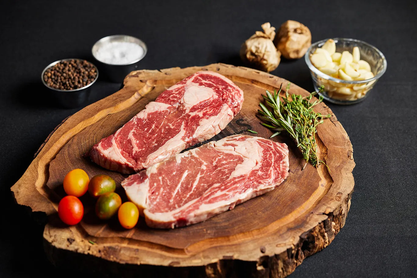 Two Delmonico Ribeye steaks on a cutting board, garnished with fresh herbs and an array of spices.