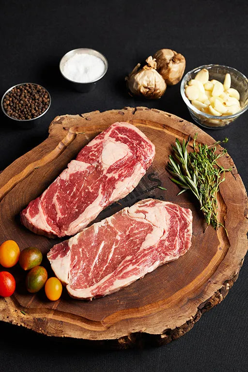 Two Delmonico Ribeye steaks on a cutting board, garnished with fresh herbs and an array of spices.