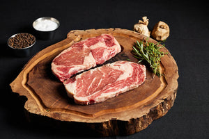 Two Delmonico Ribeye steaks on a cutting board, garnished with fresh herbs and an array of spices.