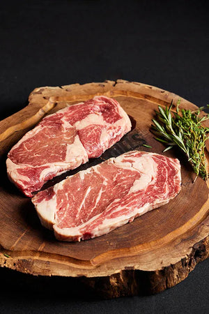 Two Delmonico Ribeye steaks on a wooden board, complemented by sprigs of rosemary.