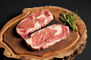 Two Delmonico Ribeye steaks on a wooden board, complemented by sprigs of rosemary.