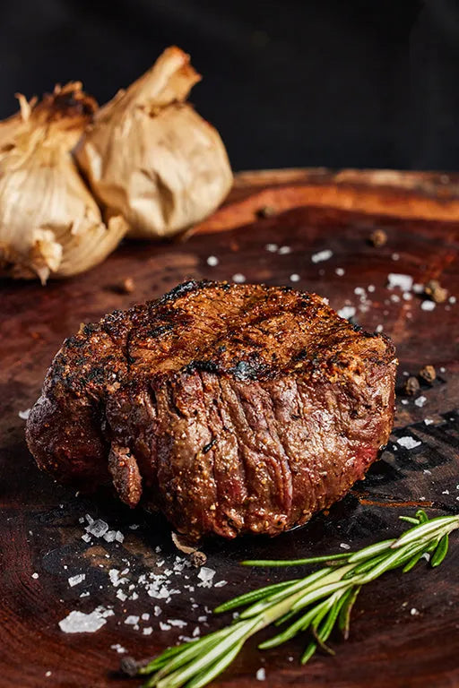 A filet mignon steak displayed on a wooden cutting board, accompanied by garlic and rosemary for flavor.