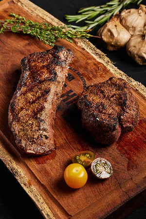 Filet Mignon and New York Strip steaks rest on a wooden cutting board, complemented by herbs and tomatoes.