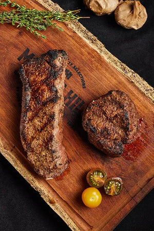 A wooden cutting board displays two grilled steaks, Filet Mignon and New York Strip, accompanied by tomatoes and herbs.