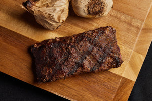 A grilled top sirloin steak resting on a cutting board, accompanied by garlic.
