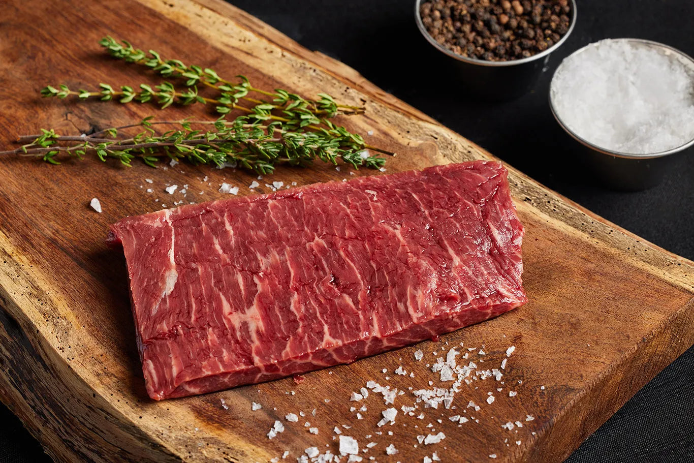 A top sirloin steak resting on a cutting board, accompanied by salt and pepper and herbs for seasoning.