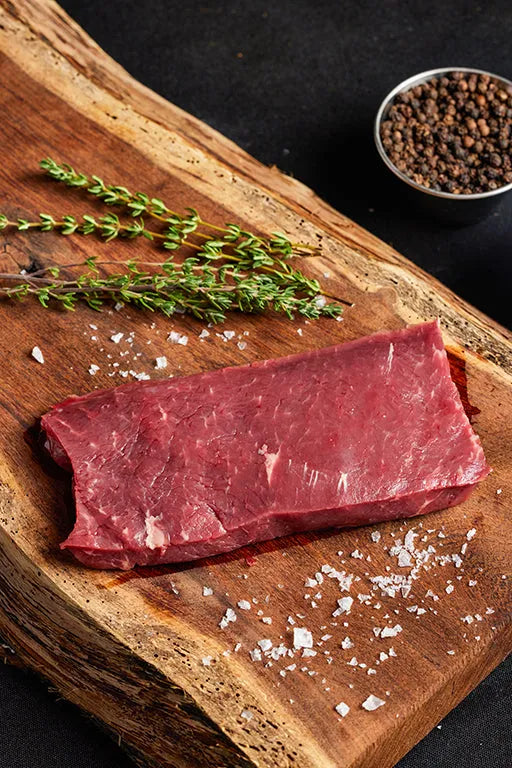 A flatiron steak on a cutting board, surrounded by an array of spices, ready for preparation and cooking.