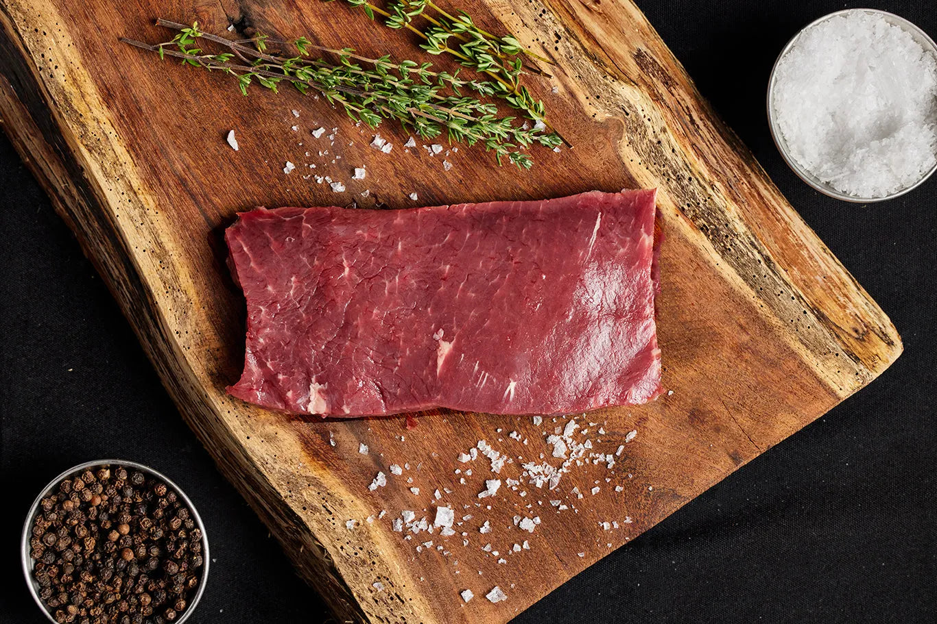 A flatiron steak on a cutting board, surrounded by an array of spices, ready for preparation and cooking.