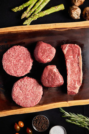 A cutting board displaying a bundle of filet mignon, New York strip, burgers, fresh vegetables, and aromatic herbs.