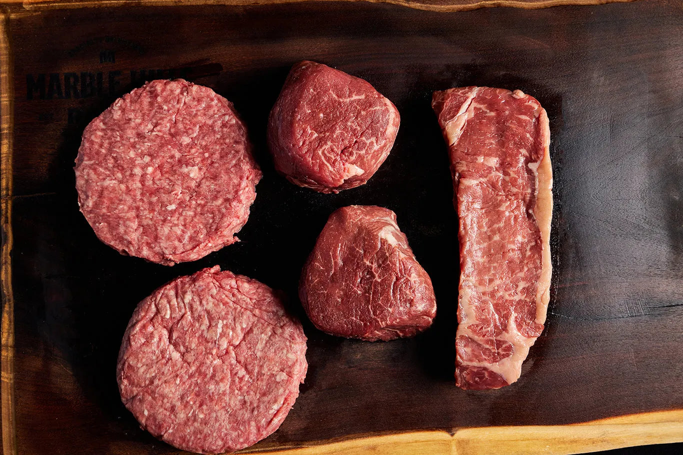 A cutting board displaying a bundle of filet mignon, New York strip and burgers.