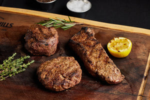 A wooden cutting board displays three steaks: filet mignon, New York strip, and burgers, garnished with lemon and herbs.