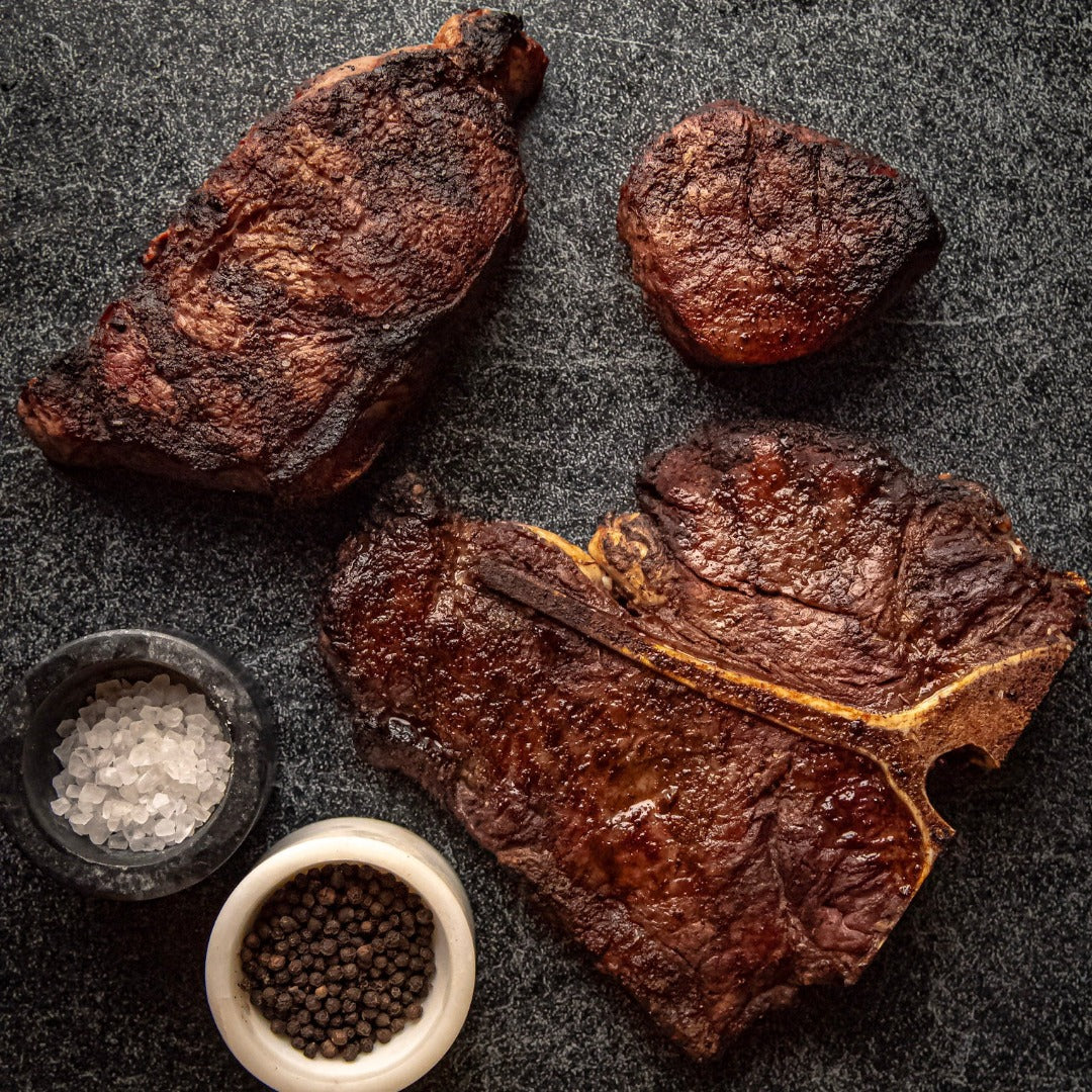 Grilled steakhouse trio - filet mignon, NY strip, and porterhouse - seasoned with salt and pepper on a black background.