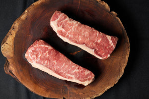 A wooden board displays two USDA Choice New York Strip steaks.