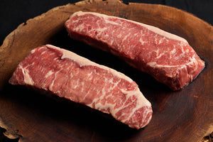 A wooden board displays two fresh USDA New York Strip steaks.