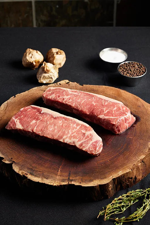 A wooden board displays two USDA New York Strip steaks, arranged with herbs and spices for flavor.