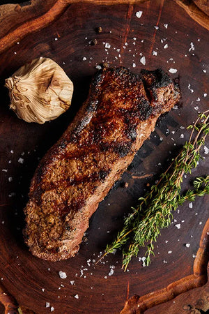 USDA Prime New York Strip steak displayed on a wooden board, accompanied by garlic and herbs for flavor.