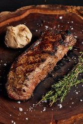 A grilled New York Strip steak on a wooden board, garnished with garlic and fresh herbs.