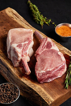 A wooden cutting board displays two pork chops, arranged with an assortment of spices.