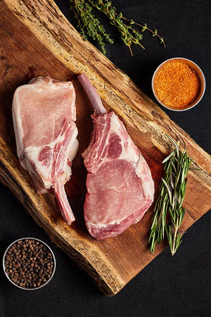 A wooden cutting board displays two pork chops, arranged with an assortment of spices.