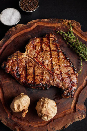 A grilled Porterhouse USDA Choice steak rests on a wooden cutting board, surrounded by herbs and spices.