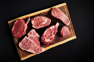 A wooden cutting board displaying a bundle of raw Filet Mignon, New York Strip, Ribeye, and Porterhouse steaks.