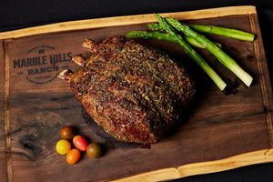 A 3-bone prime rib roast on a cutting board, accompanied by fresh asparagus and ripe tomatoes.