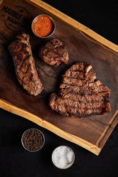 A wooden cutting board displaying a bundle of Filet Mignon, New York Strip, and Porterhouse steaks with seasoning.