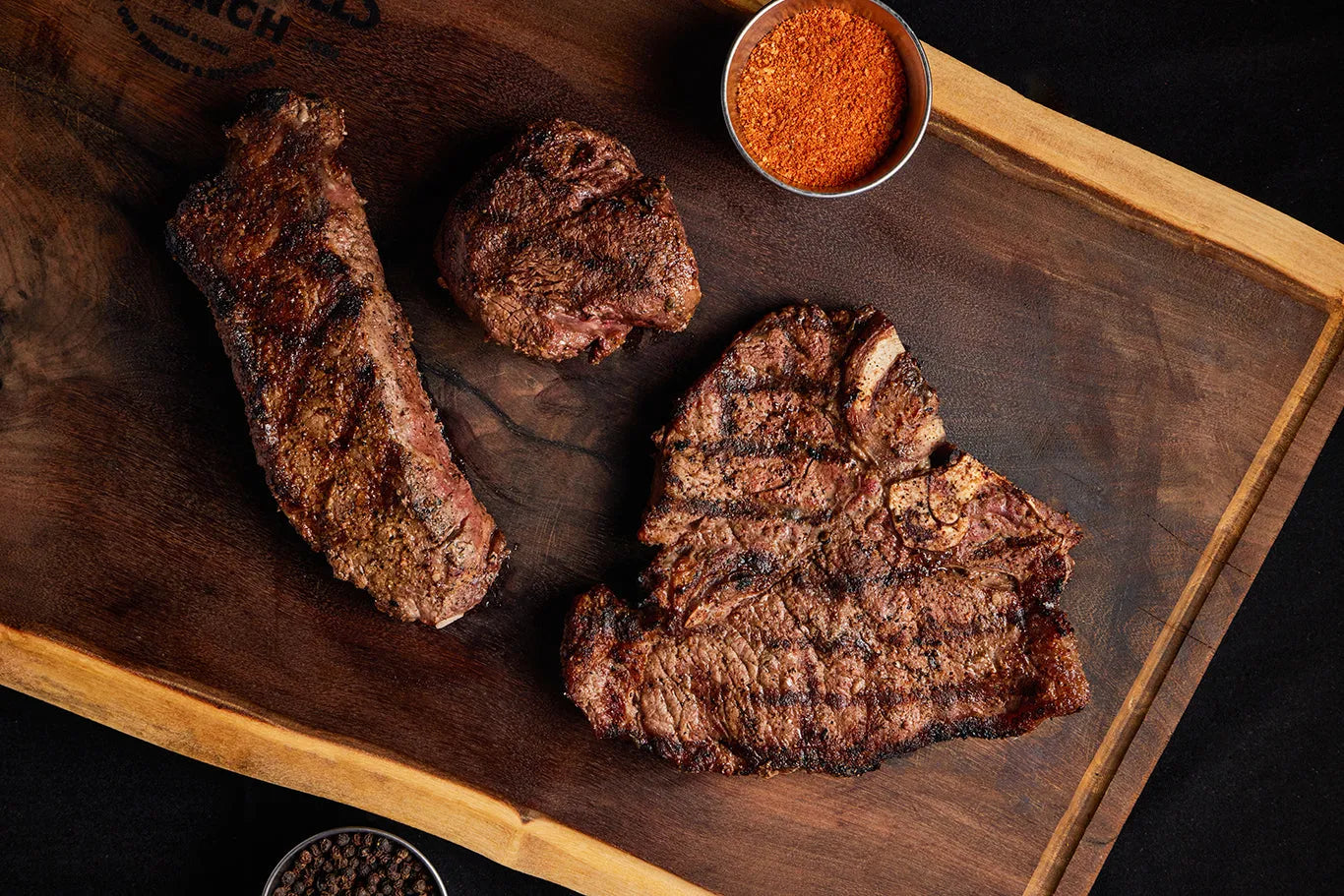 A wooden cutting board displaying a bundle of Filet Mignon, New York Strip, and Porterhouse steaks with seasoning.