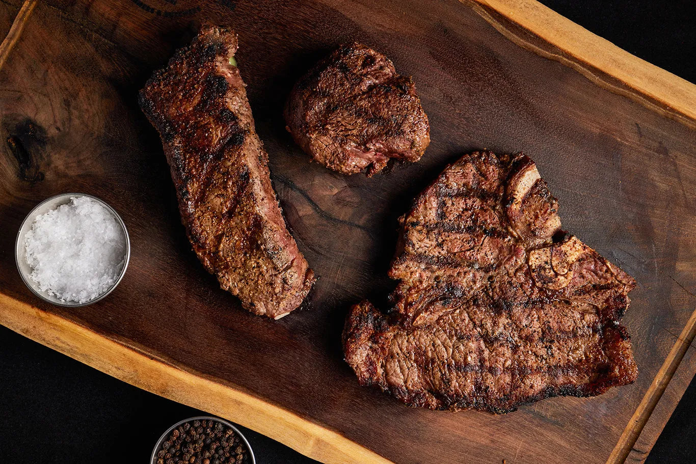 A wooden cutting board displaying a bundle of Filet Mignon, New York Strip, and Porterhouse steaks with seasoning.