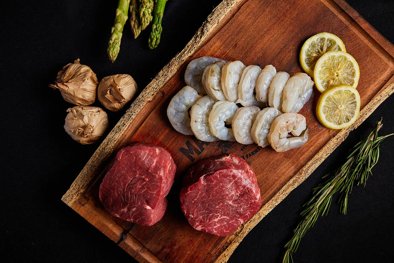 A cutting board displaying a shrimp and steak bundle, accompanied by asparagus and garlic, ready for preparation.