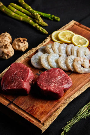 A cutting board displaying a shrimp and steak bundle, accompanied by asparagus and garlic, ready for preparation.