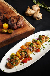 A surf and turf dish featuring steak on a wooden cutting board and shrimp on a white plate.