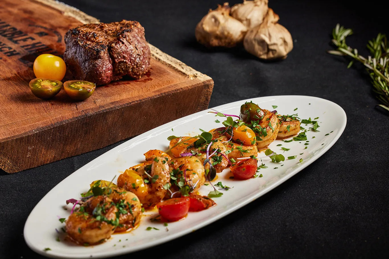 A surf and turf dish featuring steak on a wooden cutting board and shrimp on a white plate.