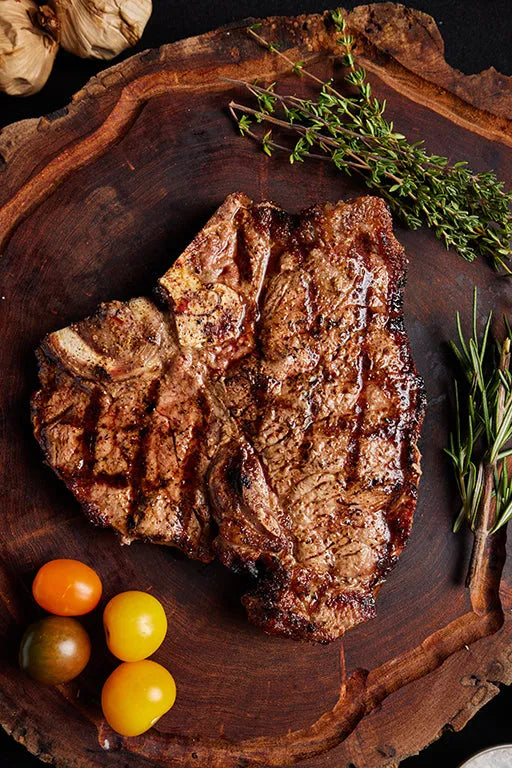 A grilled T-Bone USDA Choice steak rests on a wooden cutting board, accompanied by tomatoes and herbs.