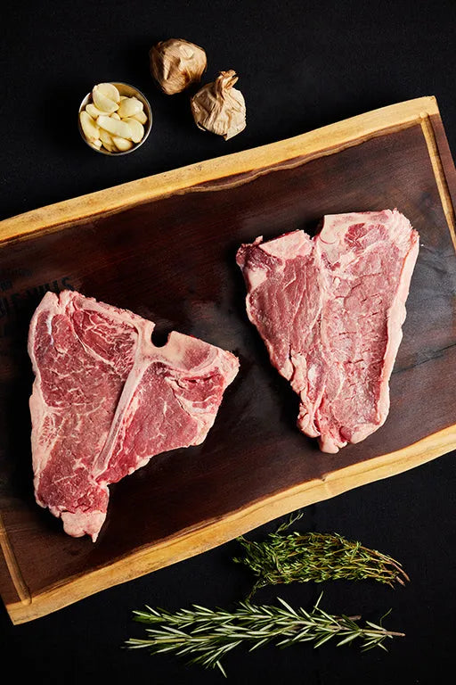 Two USDA Choice T-Bone steaks resting on a cutting board, accompanied by herbs and various spices for seasoning.