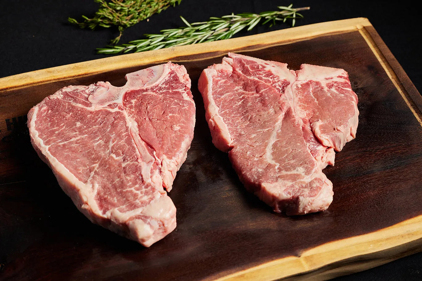 Two USDA Choice T-Bone steaks resting on a cutting board, accompanied by herbs.