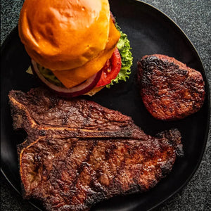 A black plate displays a T-bone steak, Filet Mignon alongside a burger, representing a meat selection.