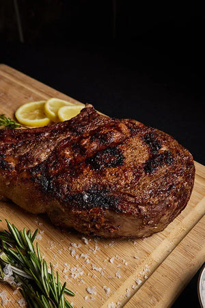 A grilled USDA Platinum Tomahawk Ribeye steak displayed on a cutting board, with lemon and and herbs for flavor.