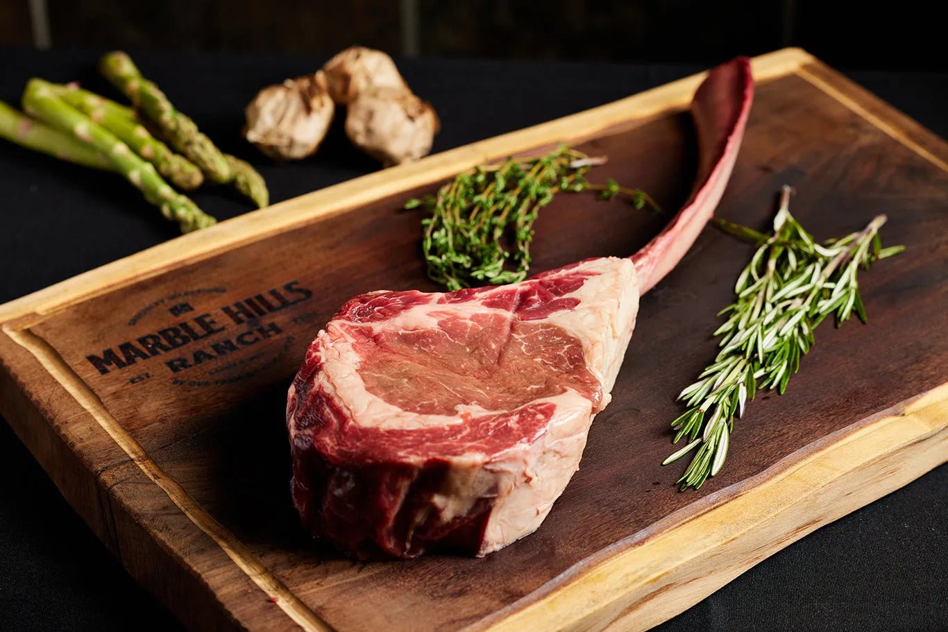 A USDA Tomahawk Ribeye steak displayed on a cutting board, accompanied by herbs and vegetables.